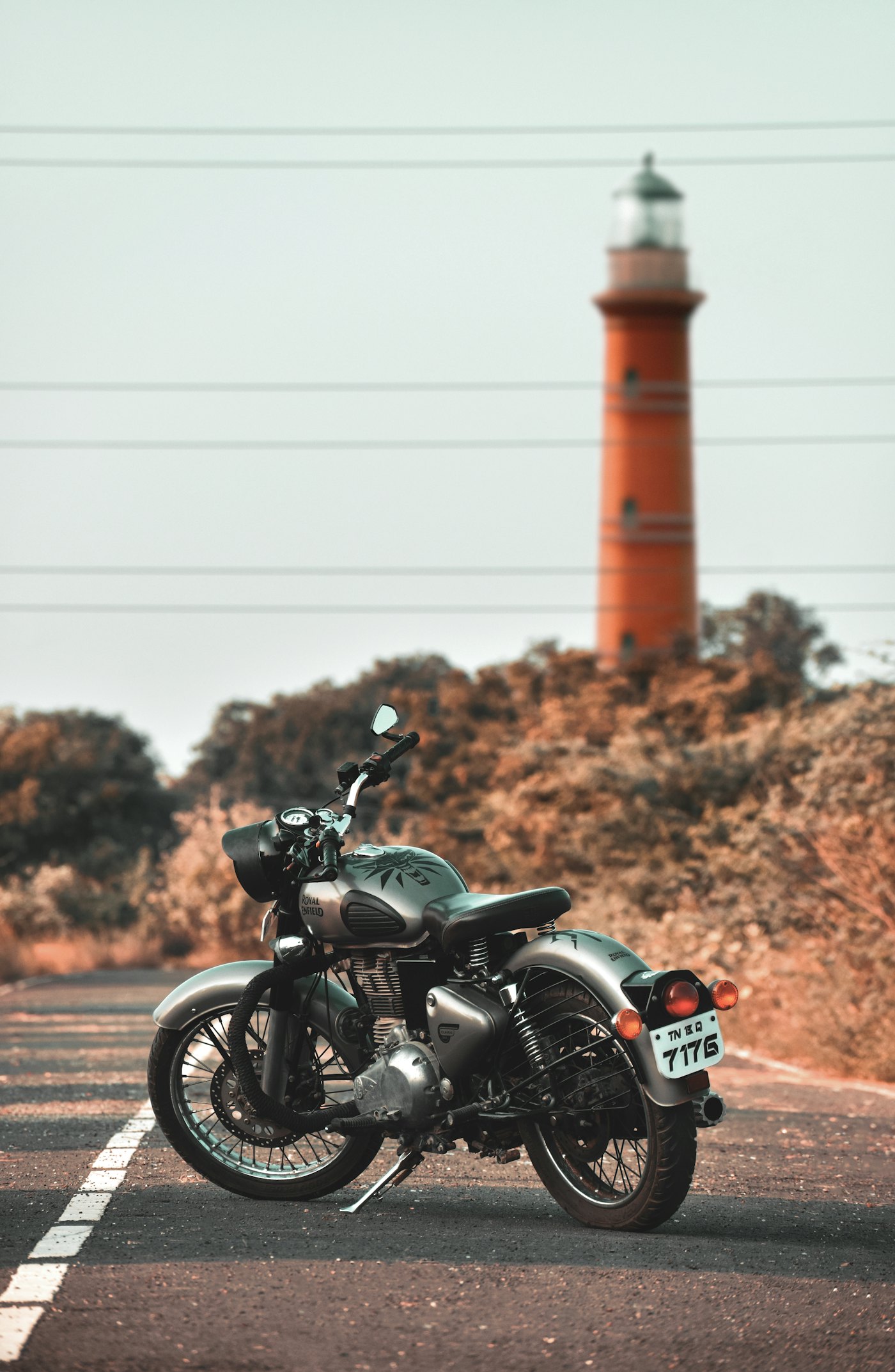 A Royal Enfield motorcycle parked on a coastal road, with a red and white lighthouse behind it
